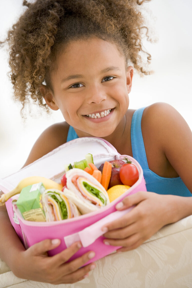 A smiling child, wearing a blue tank top, embraces their school lunch ideas with love. Their open lunchbox reveals wraps, carrots, tomatoes, a banana, and a carton drink—perfectly crafted for happy children.
