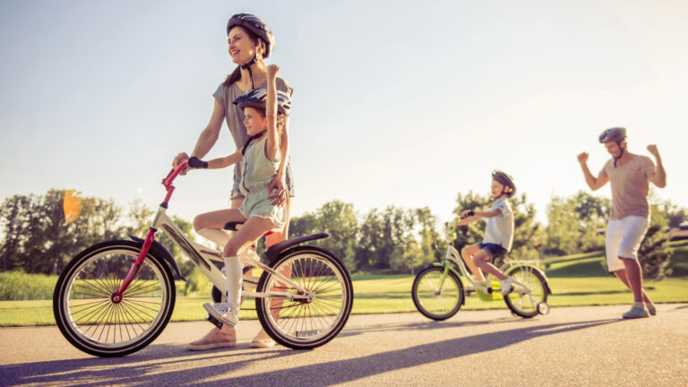 A family enjoys a spring bucket list activity, riding bicycles in the park on a sunny day. A woman and child lead on bikes, followed by another child and a man, all wearing helmets. Trees and grass form the vibrant backdrop to their family fun adventure.
