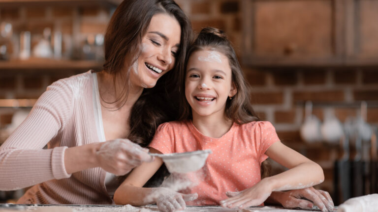 A woman and a girl are joyfully engaged in an educational baking session in the kitchen, both smiling with flour on their faces and hands. The woman is sprinkling flour while the girl eagerly stands beside her, learning that cooking can be fun for all ages.