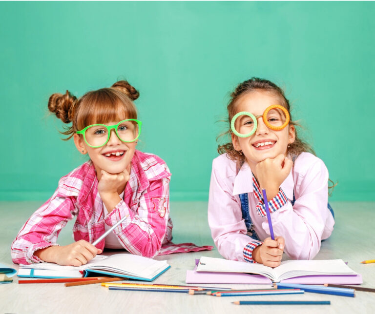 Two young girls wearing colorful glasses and plaid shirts lie on the floor, smiling with open notebooks and colored pencils in front of them.