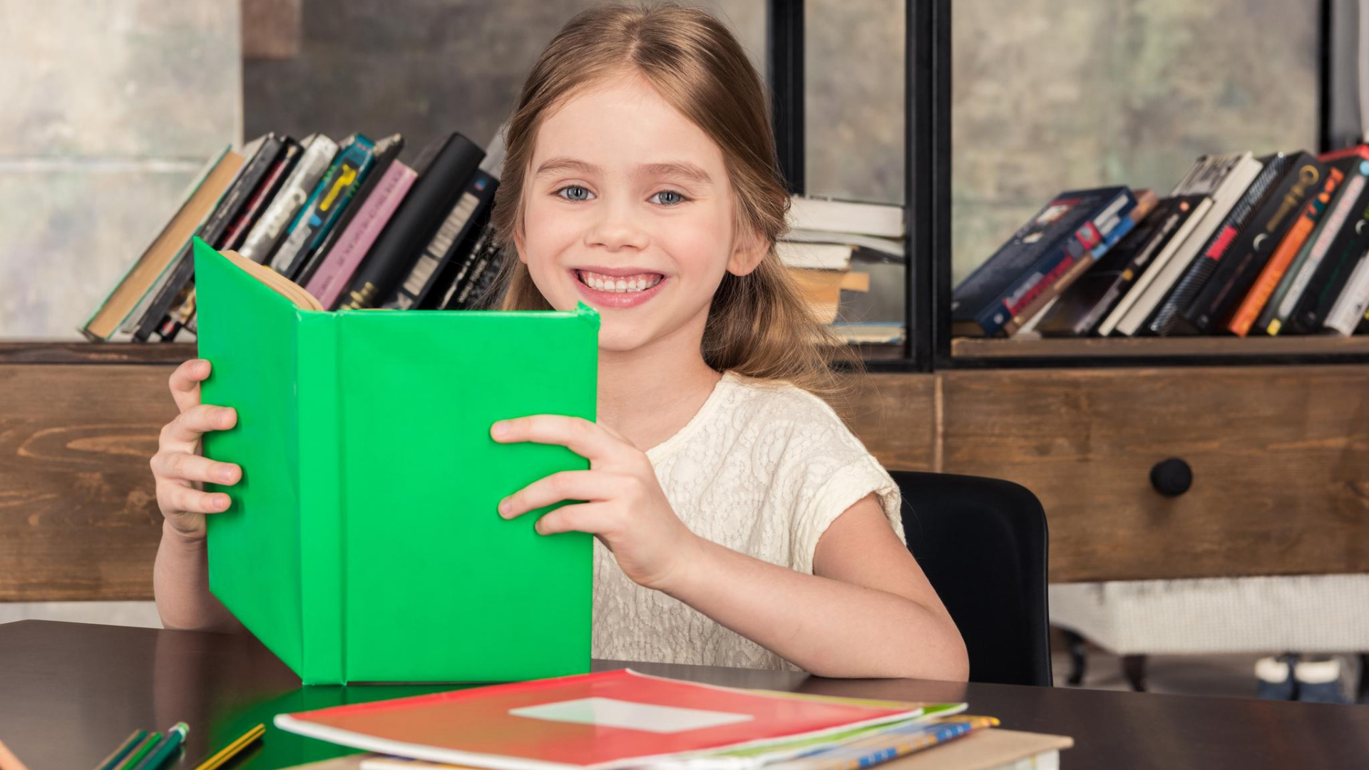 A smiling girl holds an open green book at a table, unlocking her curiosity. Behind her, a shelf of books hints at the benefits of non-fiction, especially for kids eager to explore the world.