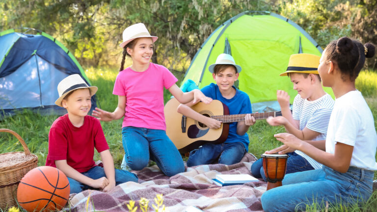 Five children sit on a blanket outdoors near tents, playing guitar and drums, smiling together at a campsite on a sunny day. A basketball and basket are beside them, perfect for fun camping activities for kids.