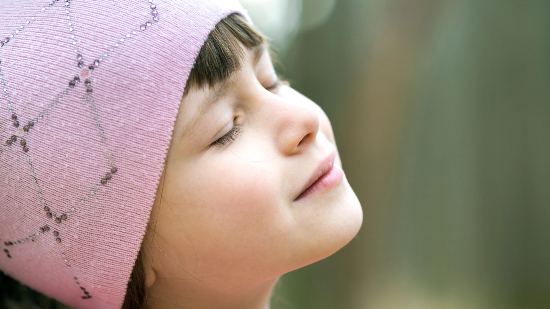 A young girl in a pink knit hat, embodying mindfulness with her eyes gently closed and a serene expression, stands outdoors against a softly blurred background.