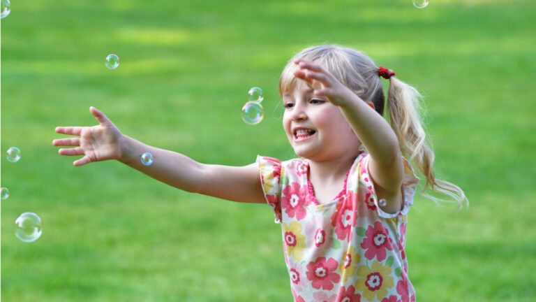 Young girl in a floral shirt with pigtails reaching out to catch bubbles while standing on green grass, enjoying classic summer fun.