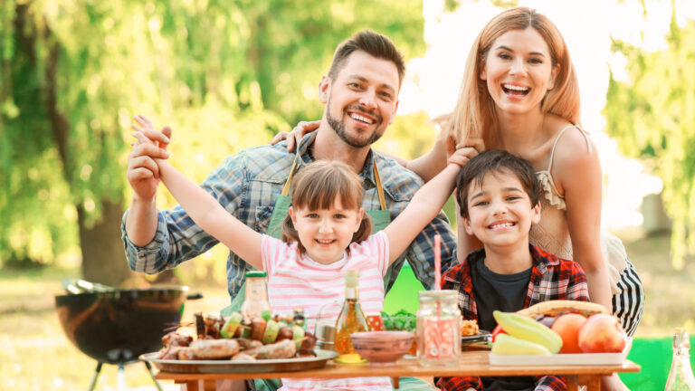 A family of four smiling at a picnic table outdoors with food and drinks, the perfect addition to their Summer Bucket List. Two children sit in front, and the parents stand behind, holding hands. A barbecue grill is visible in the background, making for the ultimate summer gathering.
