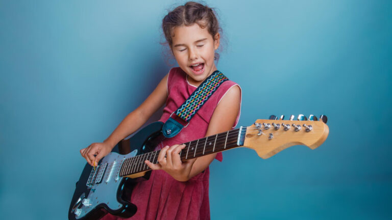 A gifted child in a red dress plays an electric guitar and sings with her eyes closed against a blue background.