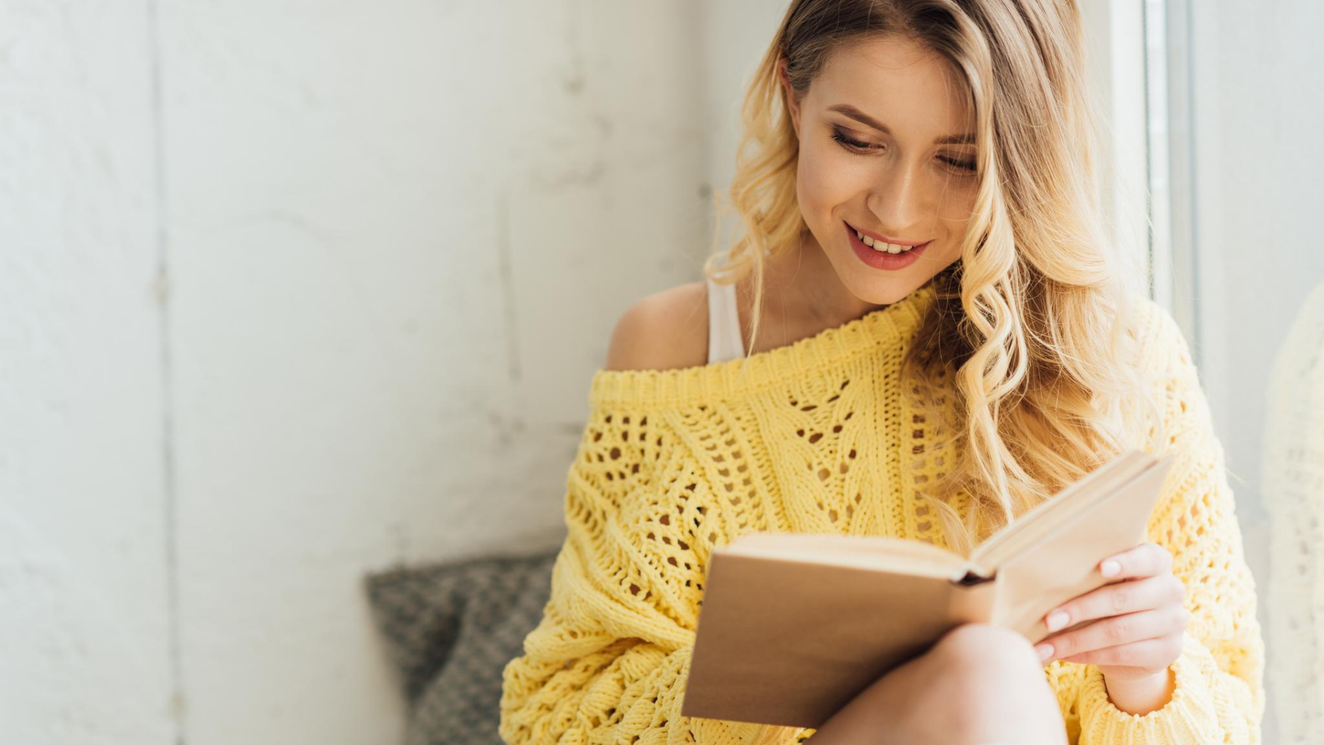 Woman with long blonde hair wearing a yellow sweater sits by a window, smiling as she enjoys one of the best self-care books—perfect summer reading for moms seeking relaxation and inspiration.