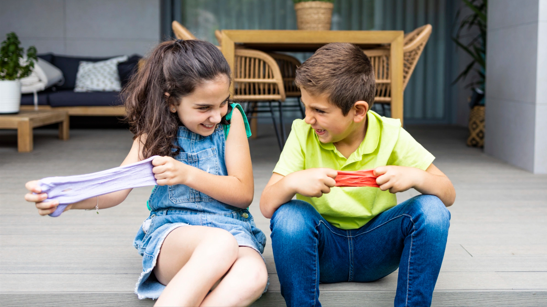 A girl and a boy sit on a porch, smiling and playing with stretchy slime—one of those boredom busting ideas that keep happy kids entertained. A wooden table and chairs are in the background.