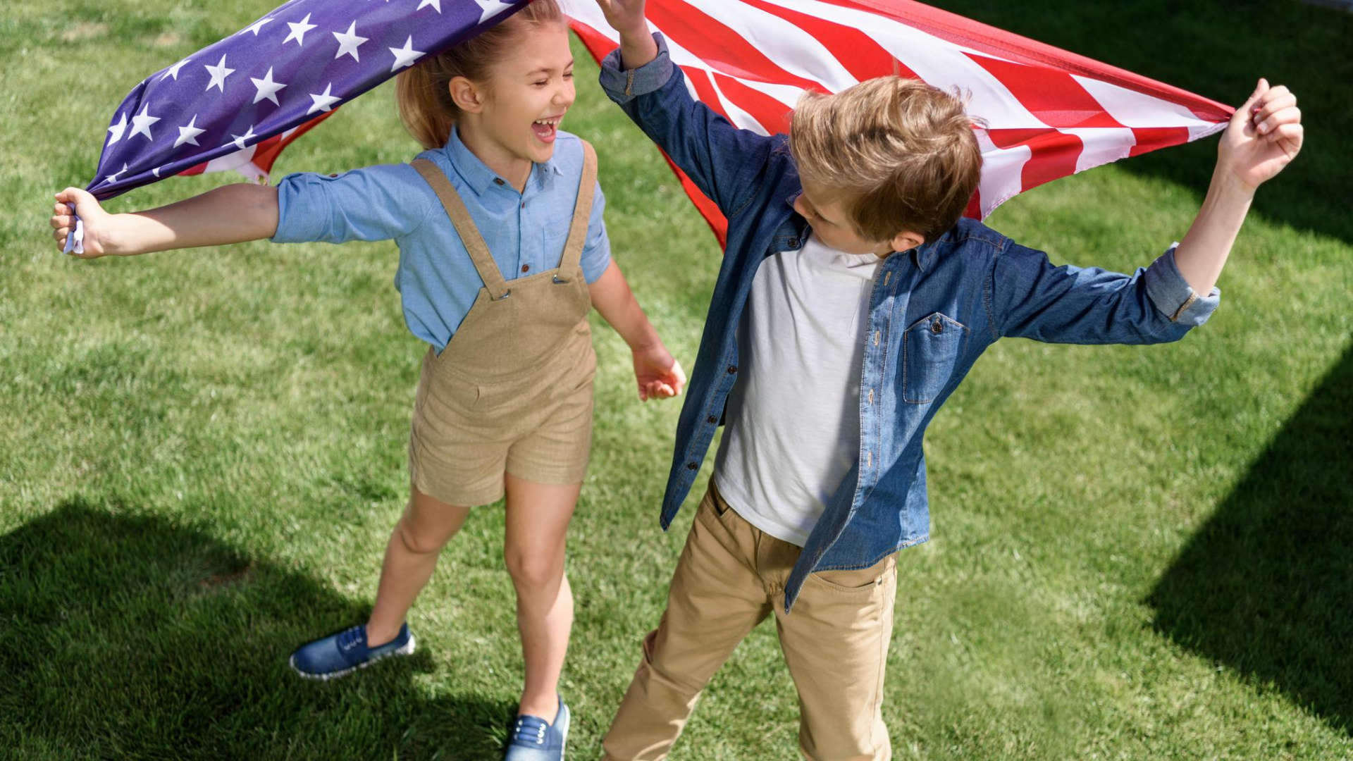 Two children stand on grass, holding an American flag above their heads, smiling at each other and sharing the fun for kids that comes with celebrating the 4th of July.