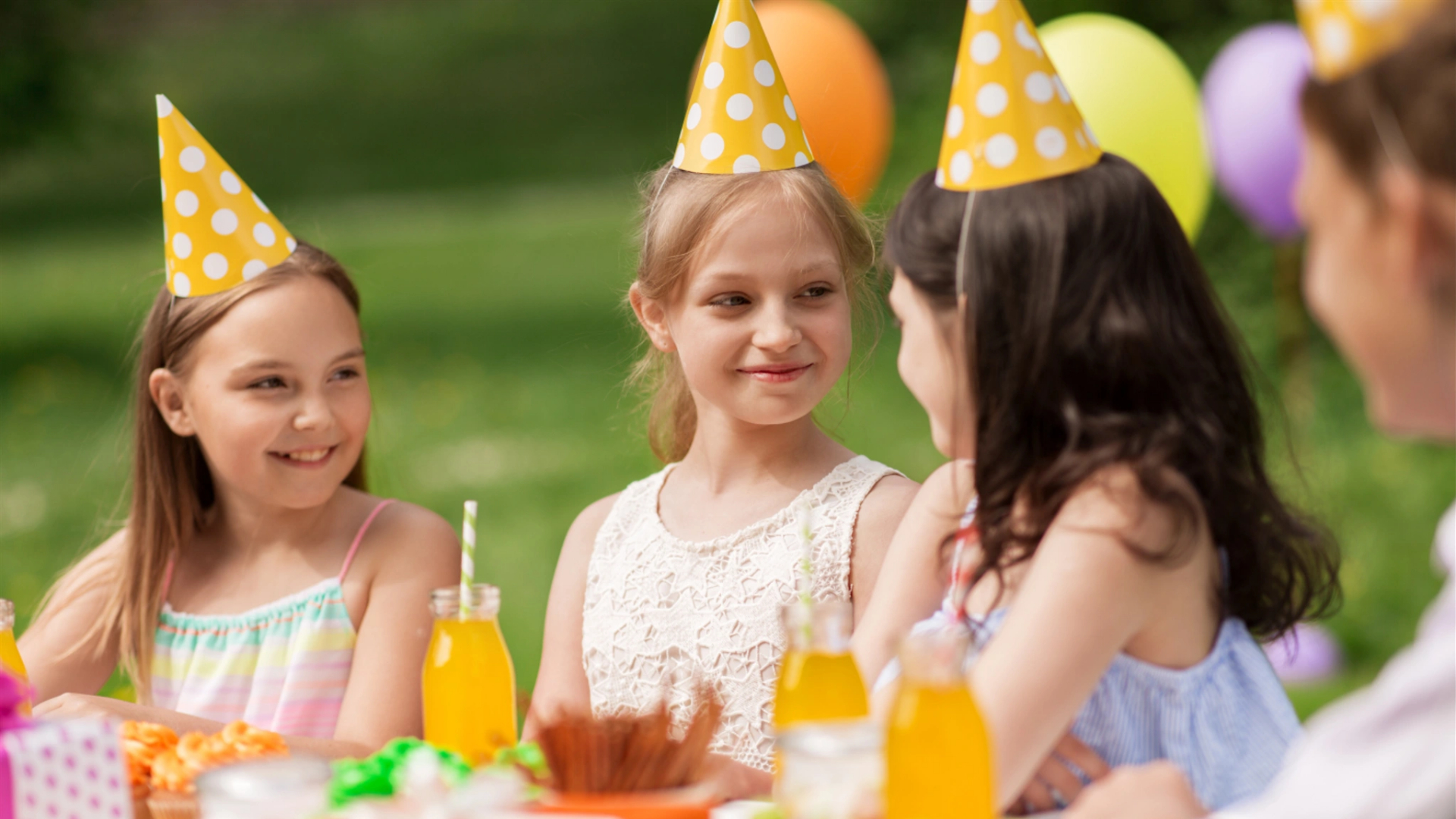Three young girls in party hats sit at an outdoor table with yellow drinks, smiling and talking amidst balloons—a joyful summer birthday celebration.