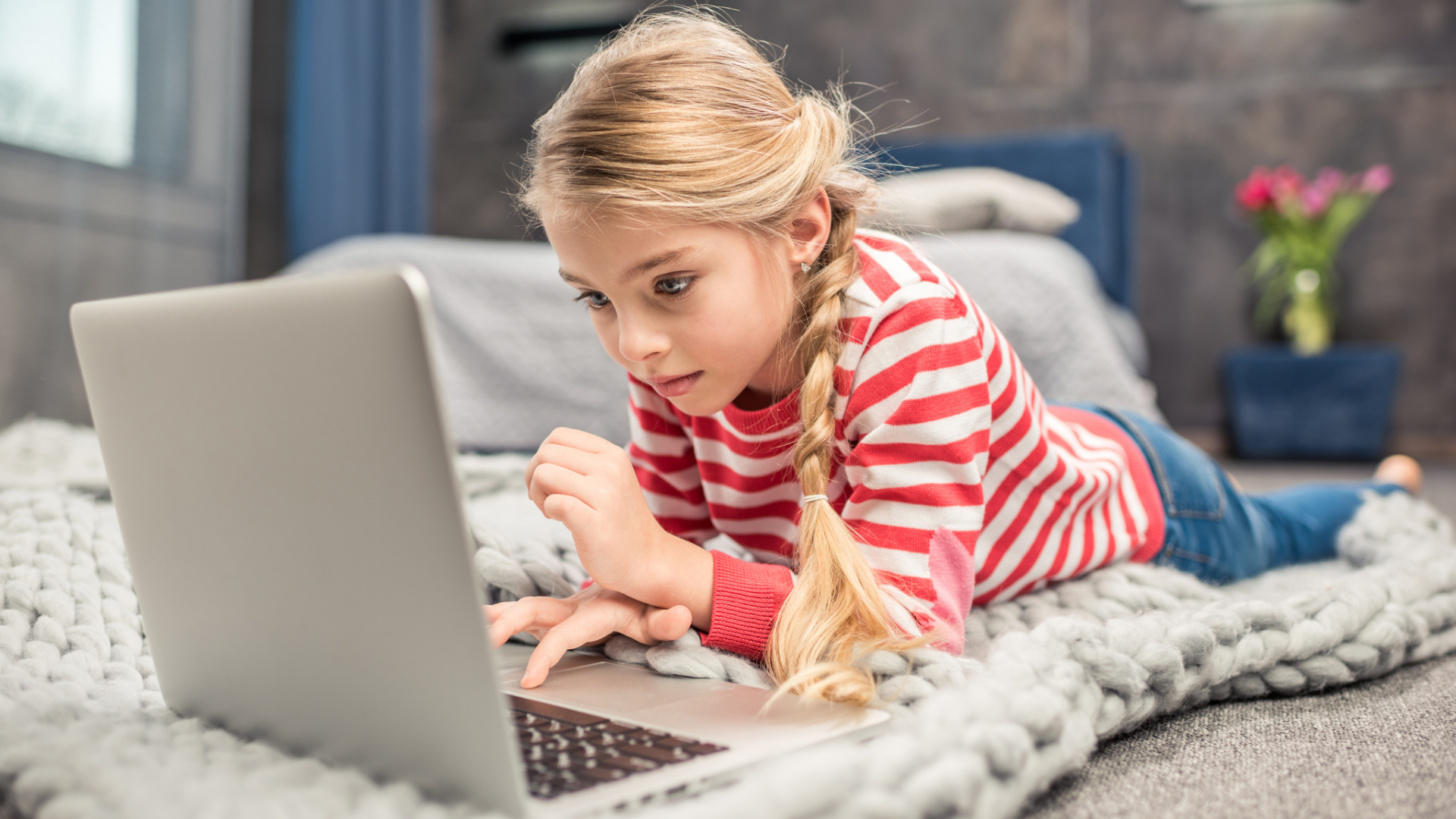 A young girl with blonde hair in a braid, wearing a red and white striped shirt, lies on a blanket using her laptop for smarter learning with AI in homeschool.