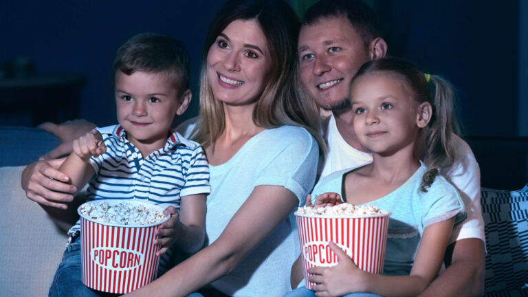 A family of four sits on a couch, smiling and holding large buckets of popcorn, enjoying a family-friendly blockbuster together in a dimly lit room.