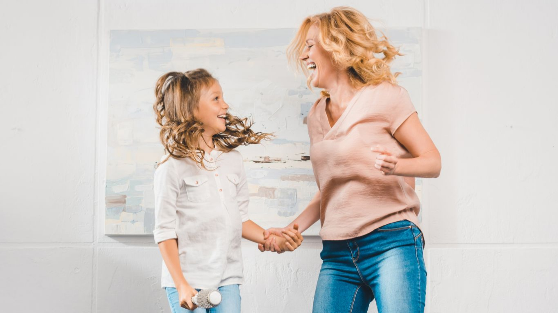 A woman and a young girl hold hands and smile while dancing together in a bright room, sharing joyful moments and encouraging each other with positive affirmations.