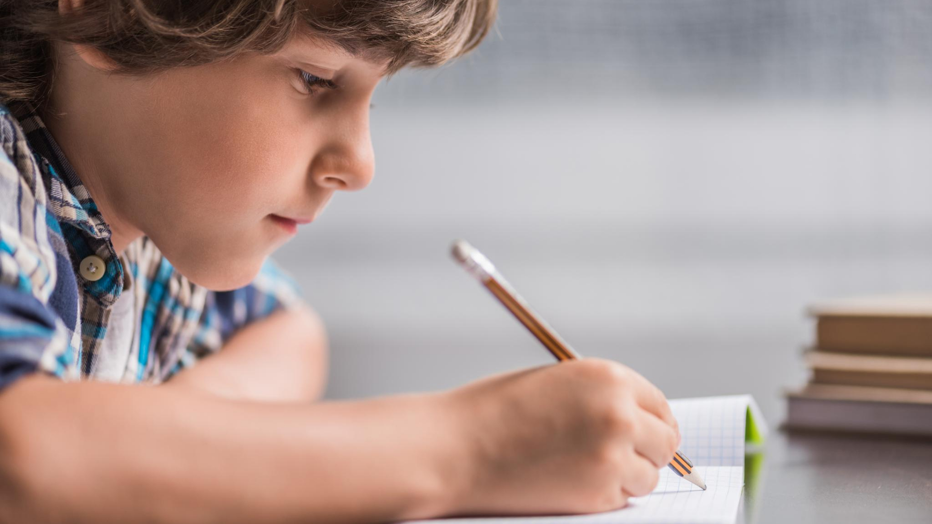 A young boy sits at a desk, writing in a notebook with a pencil. A stack of books is visible in the background, hinting at kids exploring fun fall writing prompts.