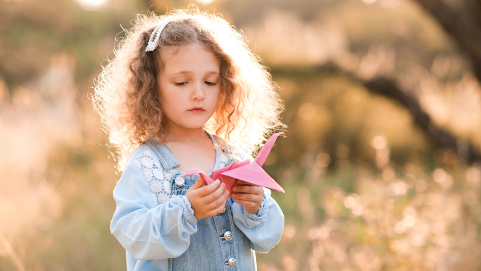 A young girl with curly hair and a blue dress stands outdoors in natural sunlight, holding an origami crane, capturing the simple joys and life lessons that kids experience every day.