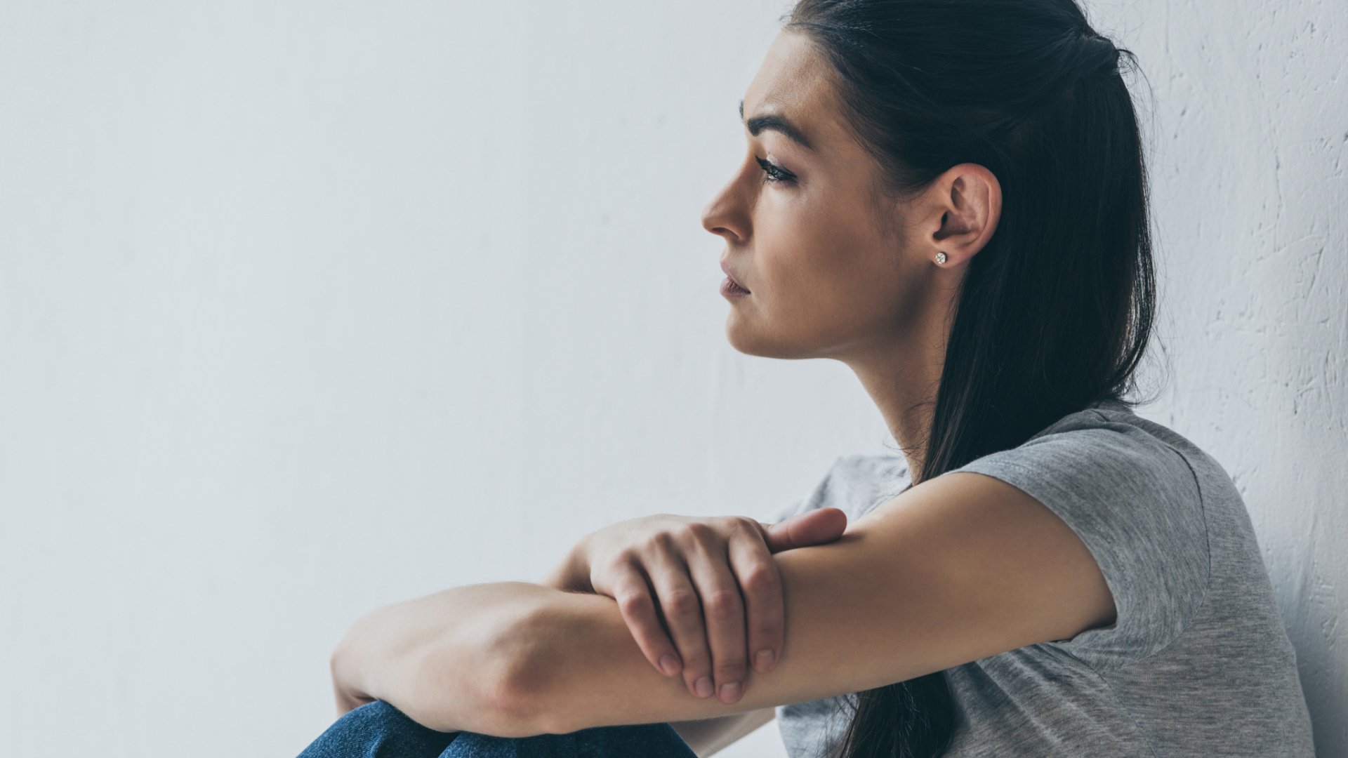 A person with long dark hair sits against a white wall, arms resting on knees, looking thoughtfully to the side—quietly holding space for sadness in a divided nation.
