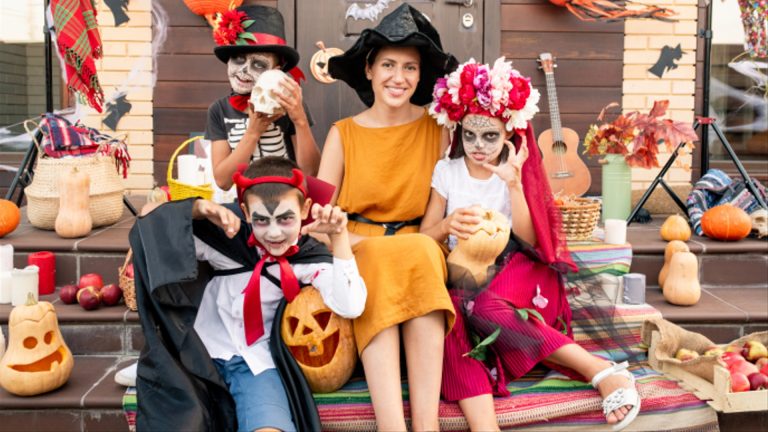 A woman and three children in Halloween costumes sit on a porch with carved pumpkins and festive decorations, surrounded by classic spooky icons—capturing the spirit of Halloween.