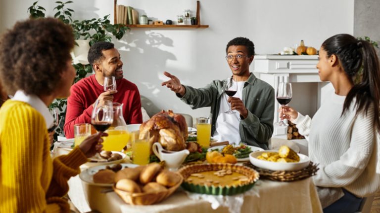 Four people sit around a dining table set with a meal, including a roast turkey and pie, holding drinks and engaging in conversation during a cozy Friendsgiving celebration.