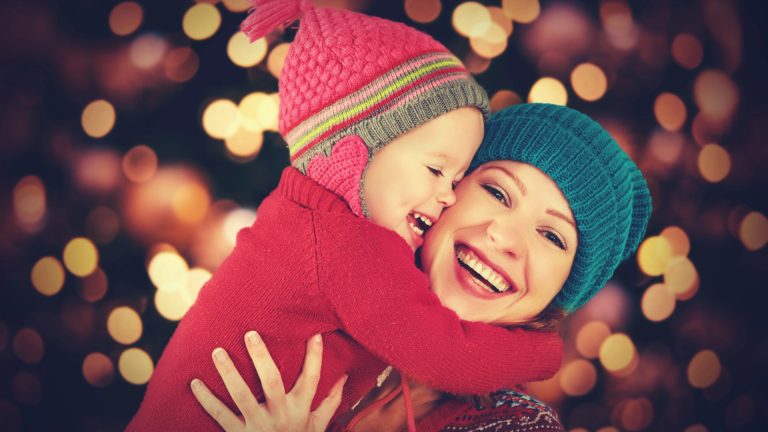 An adult and a child wearing winter hats and sweaters smile and hug each other in front of blurred festive lights, capturing the warmth of family Christmas traditions.