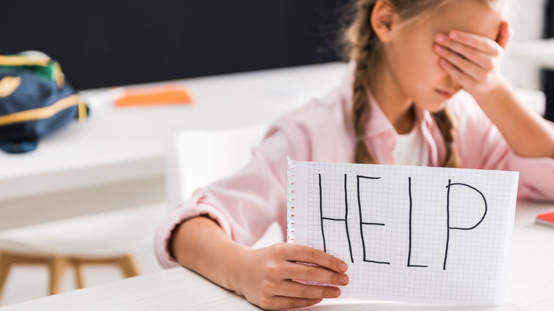A young girl sits at a desk covering her face with one hand and holding a notebook with "HELP" written in large letters, highlighting the urgent need to end bullying.