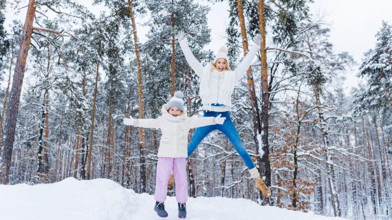 Two people dressed in winter clothing, one adult and one child, stand in a snowy forest. The adult is jumping with arms raised while the child stands with arms out, both smiling—enjoying creative fun on cold days.