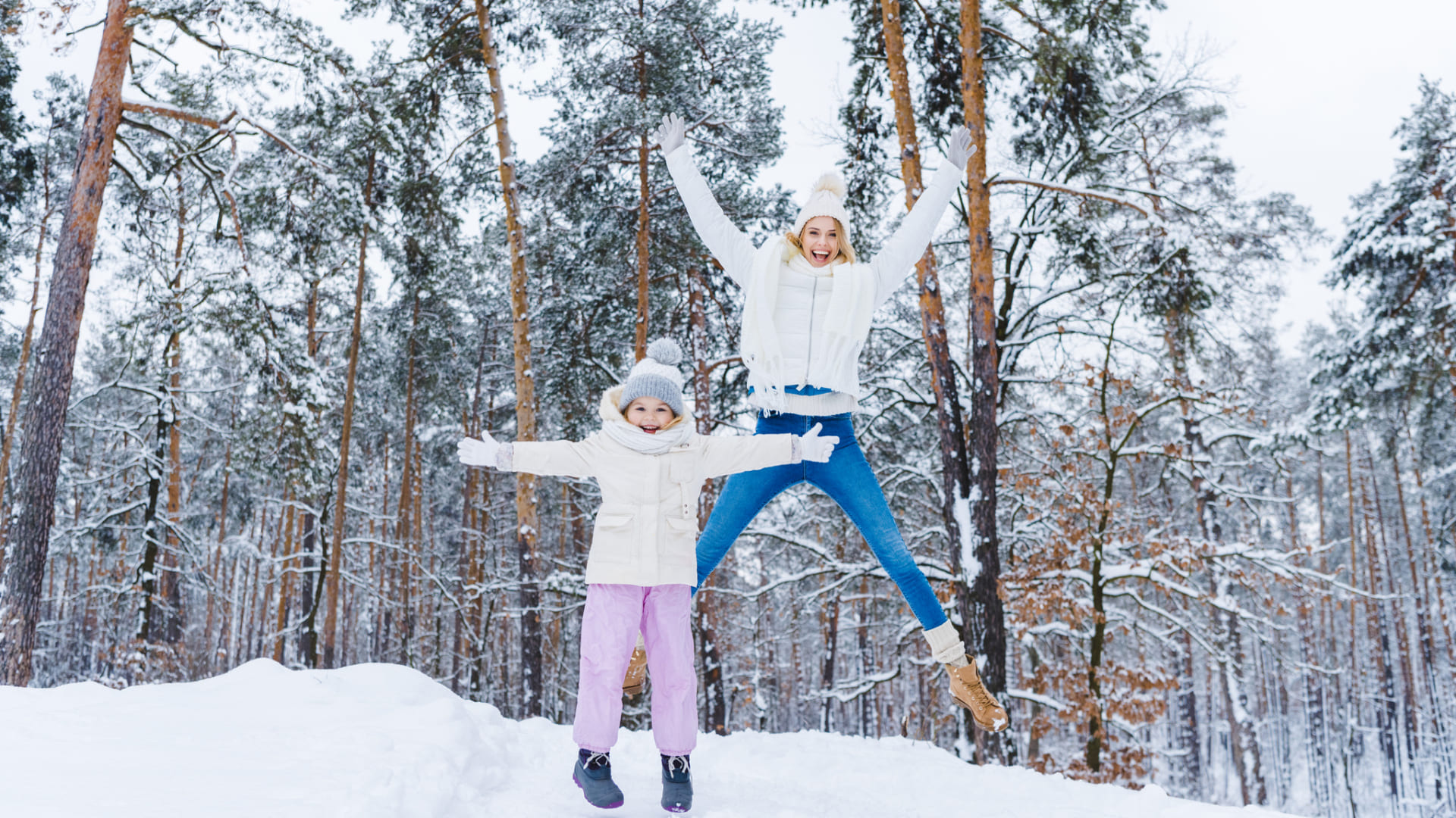 Two people dressed in winter clothing, one adult and one child, stand in a snowy forest. The adult is jumping with arms raised while the child stands with arms out, both smiling—enjoying creative fun on cold days.