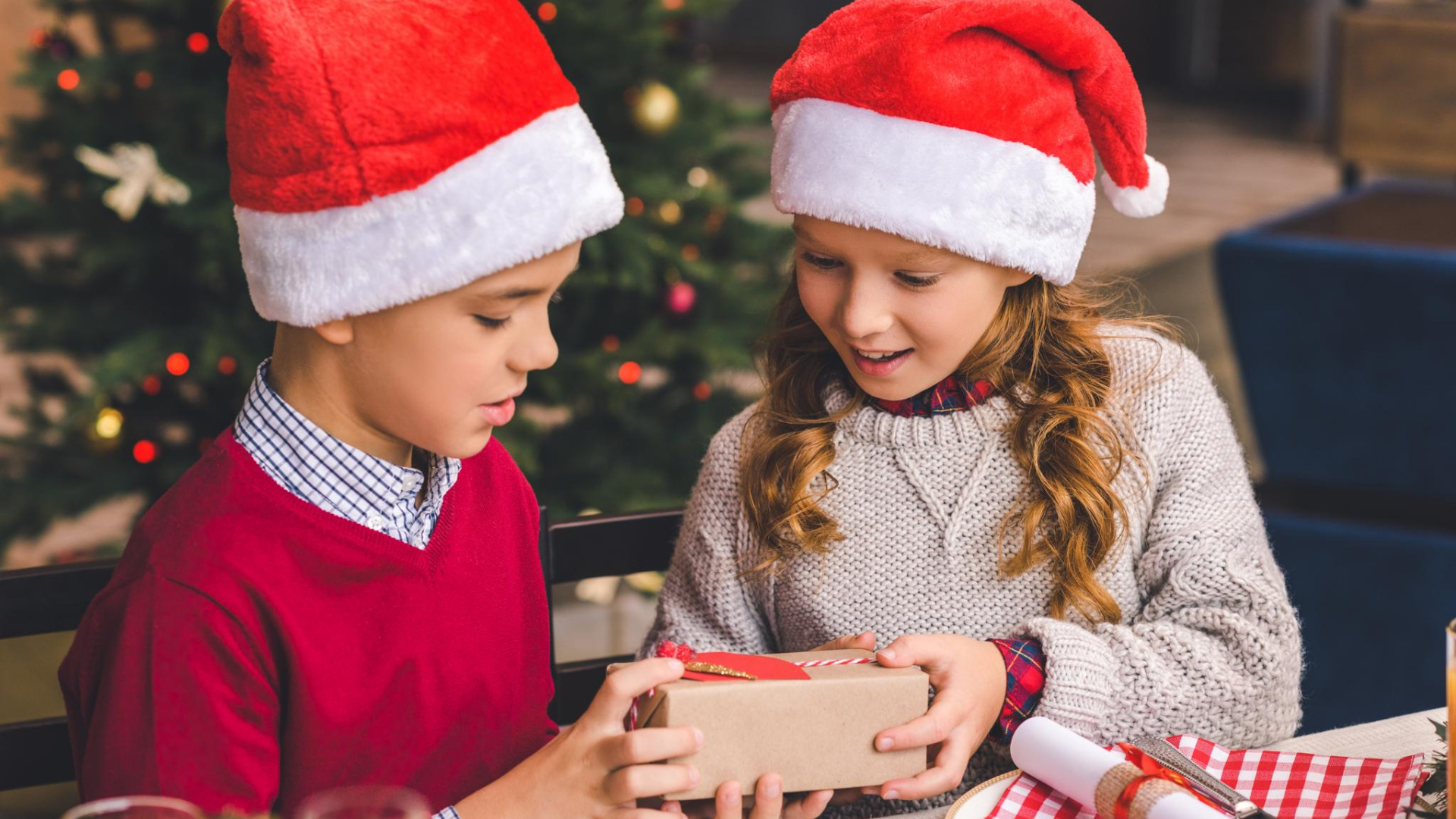 Two children wearing Santa hats sit at a table near a Christmas tree. One child gives a wrapped gift to the other, who smiles—capturing the joy of the holidays and inspiring eco-conscious kids to share and celebrate thoughtfully.