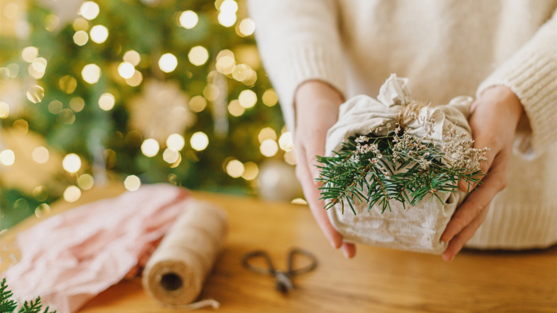 Person holding a gift wrapped in fabric with greenery and dried flowers, capturing the spirit of meaningful celebrations. A blurred Christmas tree and eco-conscious kids' wrapping materials set the festive holiday scene in the background.