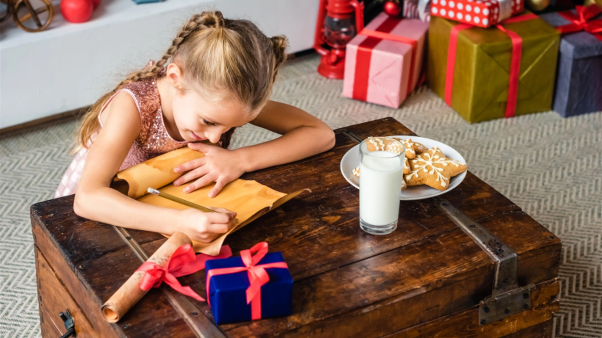 A young girl explores creative writing as she pens a letter on an envelope at a wooden table with cookies, milk, and wrapped gifts in the background.