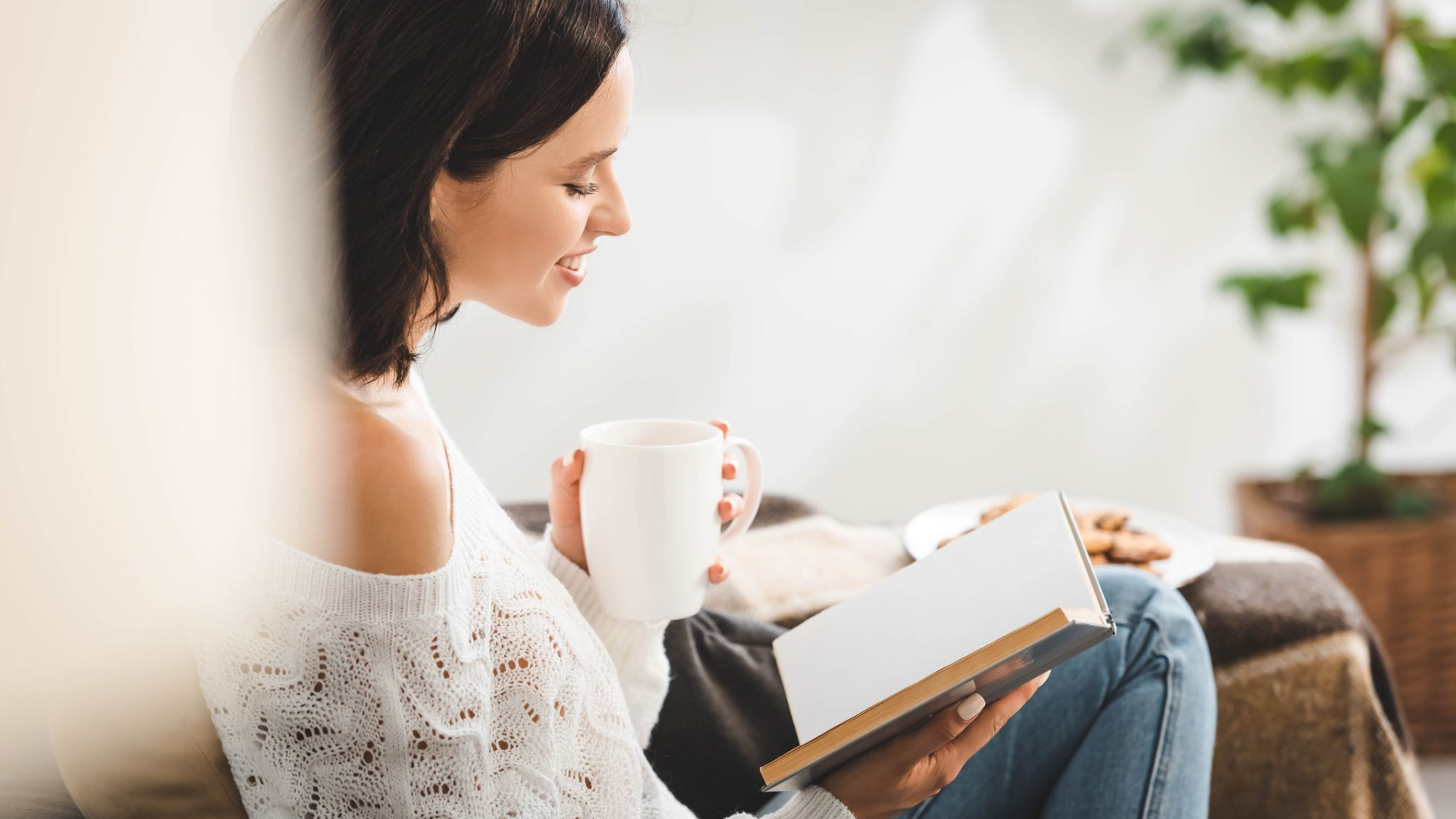 A woman sits on a couch holding a white mug and reading a book—embracing mindfulness for moms—with a plate of cookies and a plant adding coziness in the background.