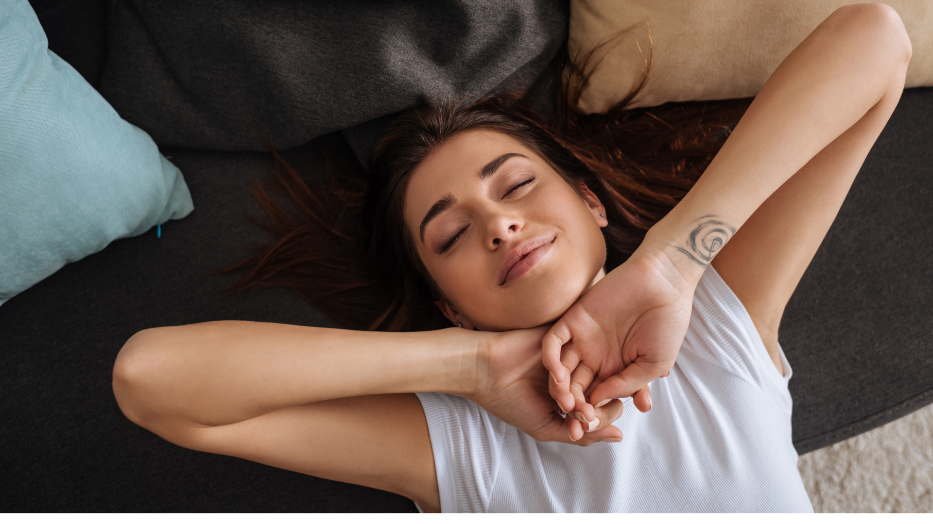 Woman lying on a couch, eyes closed, arms stretched above her head, appearing relaxed—a quiet moment of mindfulness for moms. She is wearing a sleeveless top and has a tattoo on her wrist.
