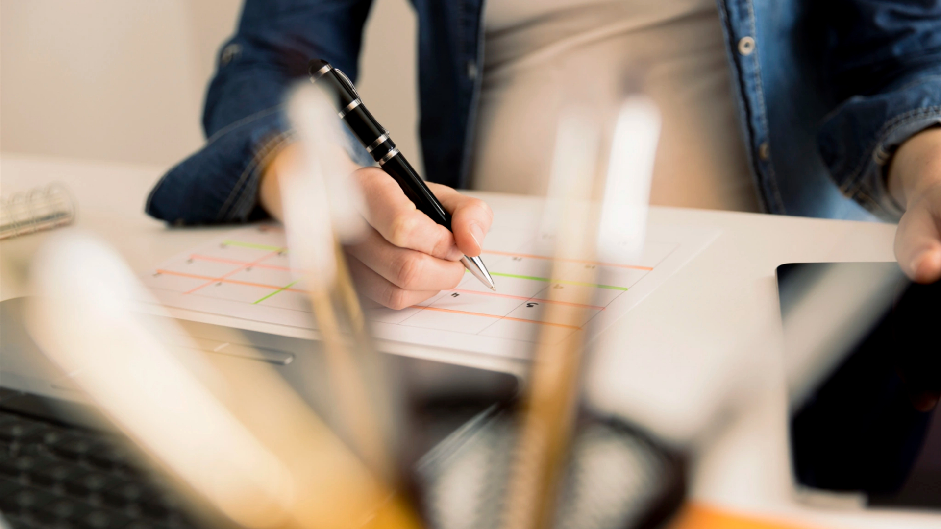 A person writes on a color-coded chart or planner at a desk, with pens and office supplies blurred in the foreground—perfect for moms seeking smart organization tips.