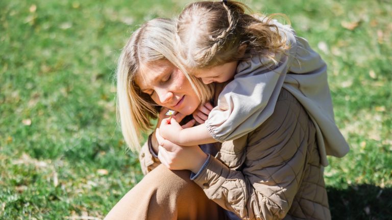 A young child hugs a woman from behind as they sit on the grass outdoors, both appearing relaxed and content in the sunlight, beautifully showing love through this gentle moment of parenting.