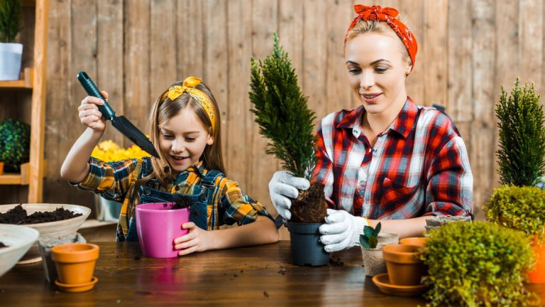 A woman and a young girl are potting small plants together at a wooden table, both wearing plaid shirts and headbands—creating cherished family memories with stay at home spring break activities.