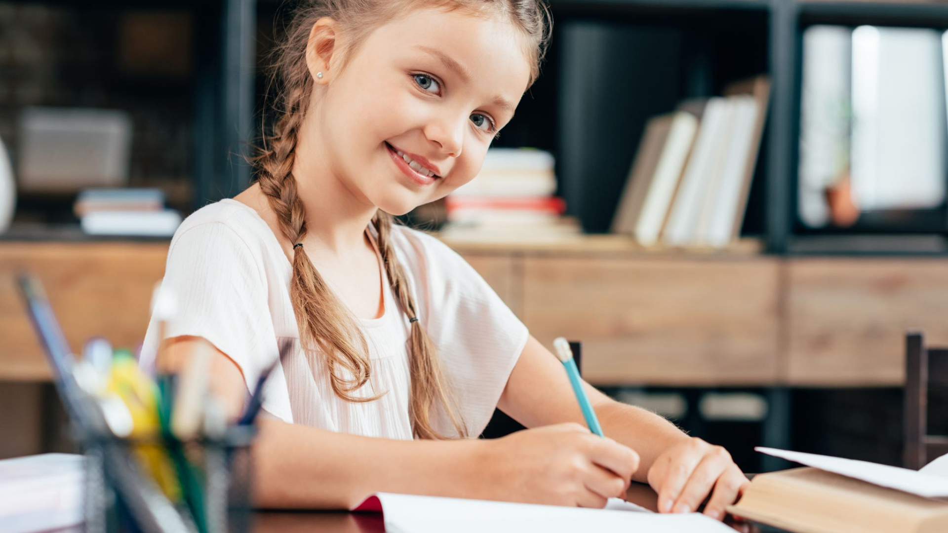 Young girl with braided hair sits at a desk, smiling at the camera while writing in a notebook filled with creative writing ideas, with shelves of books in the background.