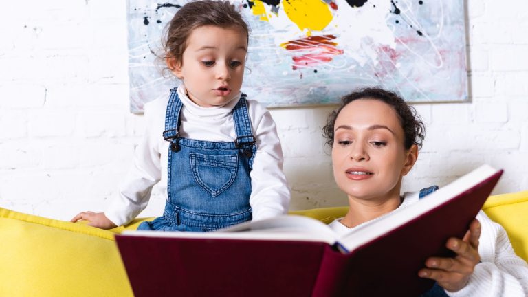 Woman reading a large book to a young girl sitting beside her on a yellow couch, surrounded by abstract artwork—an inspiring moment that shows how reading activities for kids can help your child read and foster a love of books.