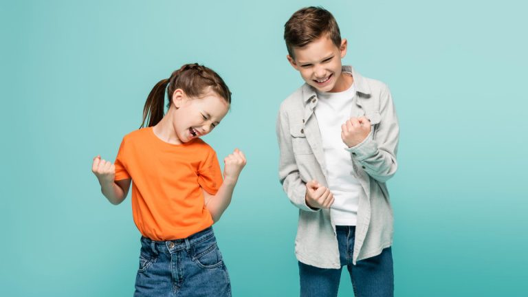 Two children standing against a blue background, smiling and clenching their fists in excitement as they build confidence and celebrate learning important life skills.