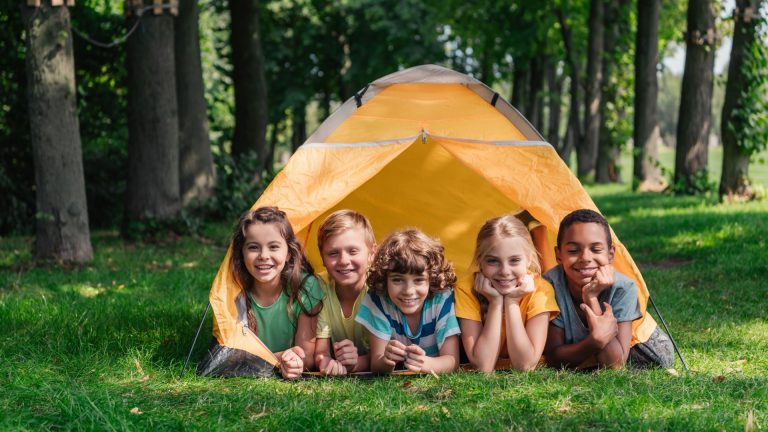 Five children lying on grass, smiling, in front of a yellow tent in a wooded area on a sunny day—perfect for stress-free camping and making the most of family camping adventures.