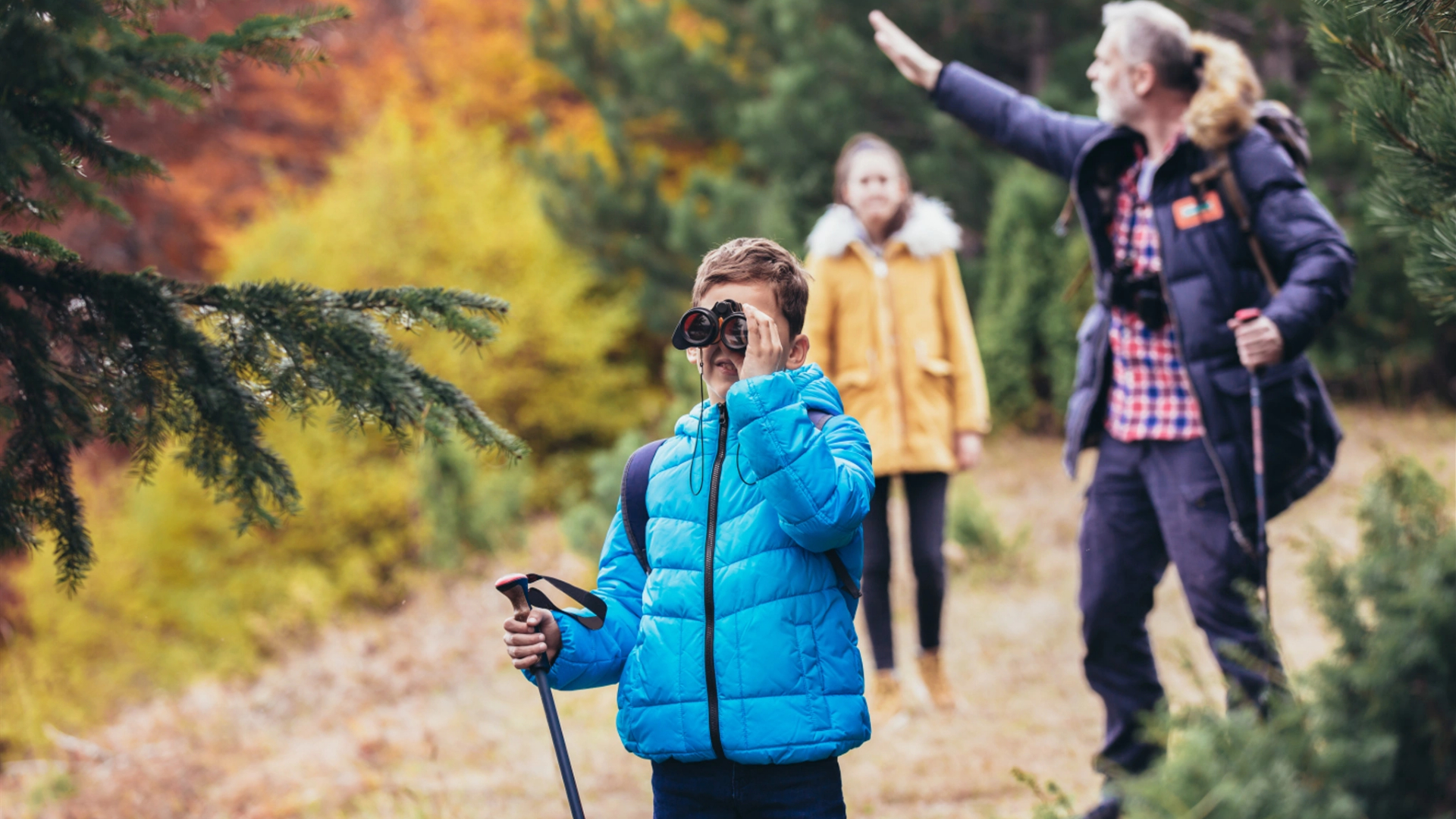 A boy in a blue jacket uses binoculars while hiking in a forest, exploring parks with kids as two adults follow behind him among vibrant autumn foliage.