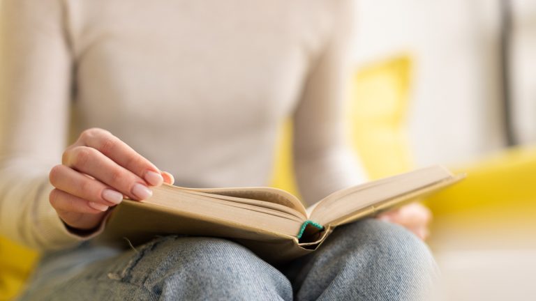 A person sits cross-legged wearing jeans and a light sweater, holding an open book—A Daughter of Fair Verona by Shakespeare—and turning a page as they begin their review.