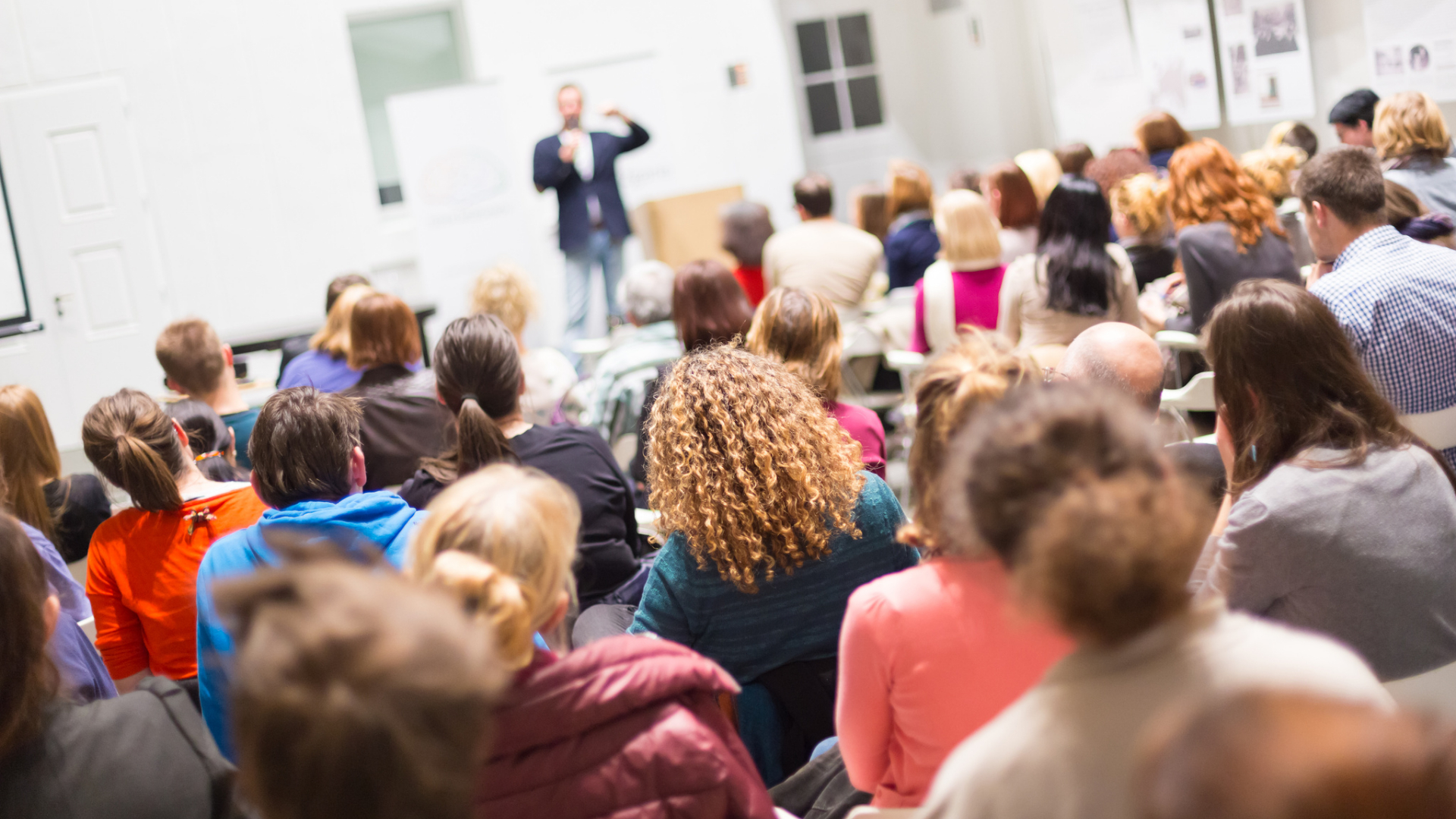 A group of people seated in a classroom or conference room attends a homeschool convention, listening to a presenter speak about homeschooling at the front of the room.