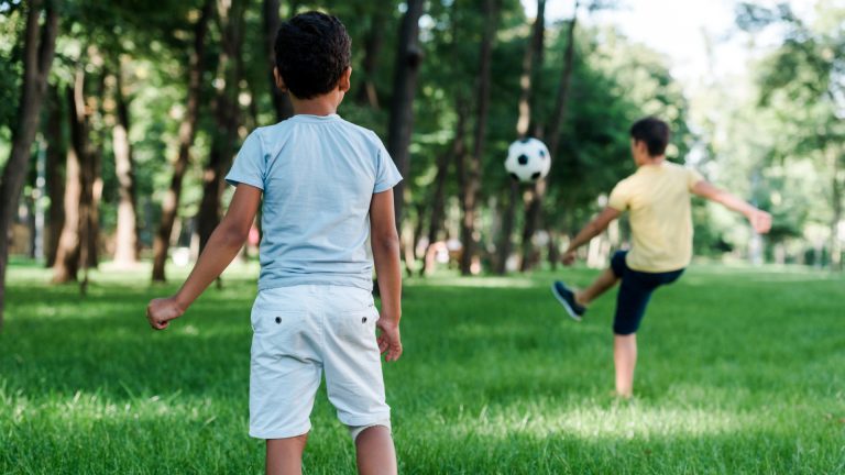 Two boys are playing soccer in a grassy park, with one boy kicking a ball while the other stands facing him. Enjoying this fun summer activity is a great way to prevent summer slide and keep active outdoors. Trees and greenery are visible in the background.