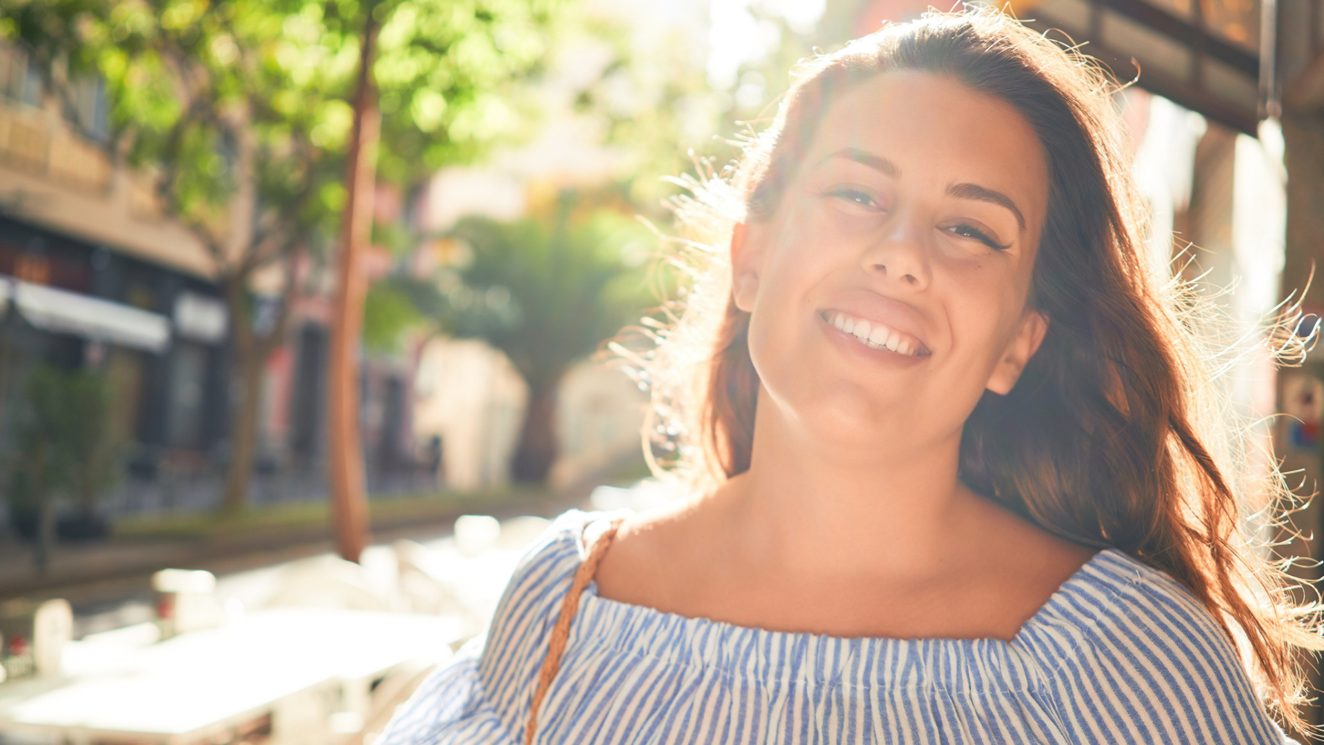A woman in a blue and white striped top smiles outdoors on a sunny day, embodying summer fun for moms, with trees and buildings blurred in the background.